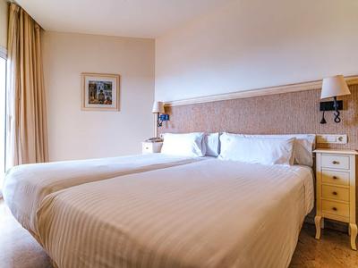 Bright hotel room with two single beds, nightstands, and wall lamps.