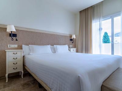 Bright hotel room with large double bed, nightstand, and curtains by the window.