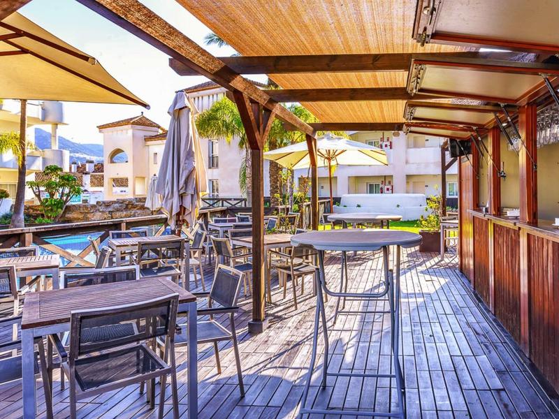 Covered terrace with wooden flooring and bar tables next to a hotel pool.