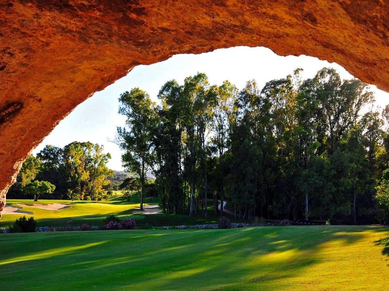 Green golf course surrounded by trees, viewed through a natural rock arch.