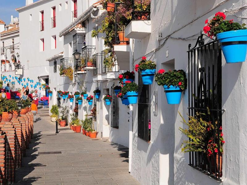 Narrow street with white houses and blue flower pots on the wall in sunny weather.