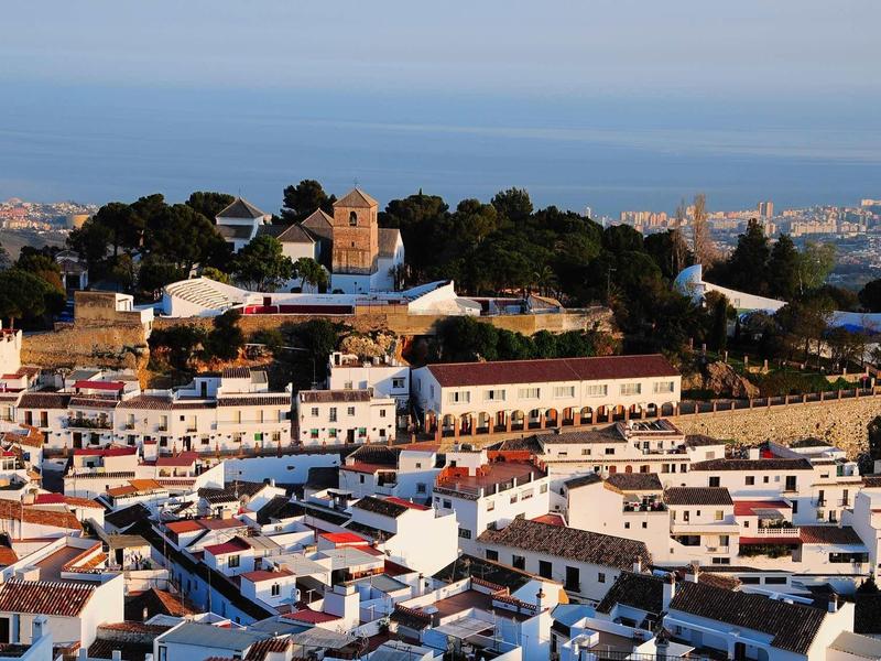 View of a town with white buildings, a historic structure on a wooded hill, and the sea in the background.