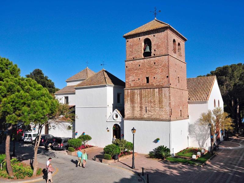 Church with brown tower and white walls under clear blue sky, surrounded by trees.