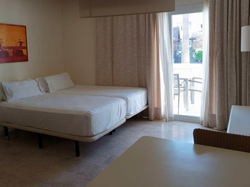 Bright hotel room with large bed, desk, TV, and balcony with seating.