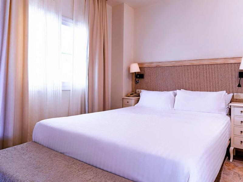 Modern bedroom with large bed, white pillows, nightstands, and softly filtered daylight.