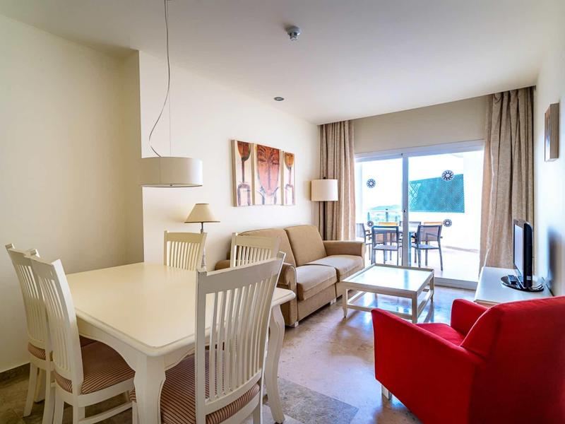 Bright living room with dining table, sofa, red armchair, and balcony overlooking the sea.