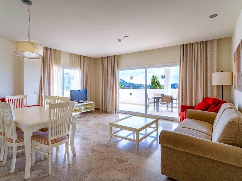 Bright living room with dining table, sofa, TV, and large window overlooking terrace and water.