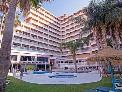 Multi-story hotel with outdoor pool and straw sun umbrellas in the garden area.