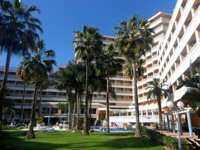 Large hotel with many palm trees and a pool area on a sunny day.