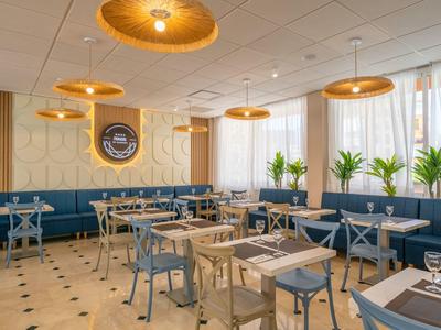 Modern restaurant with blue chairs, wooden tables, and large windows with plants.