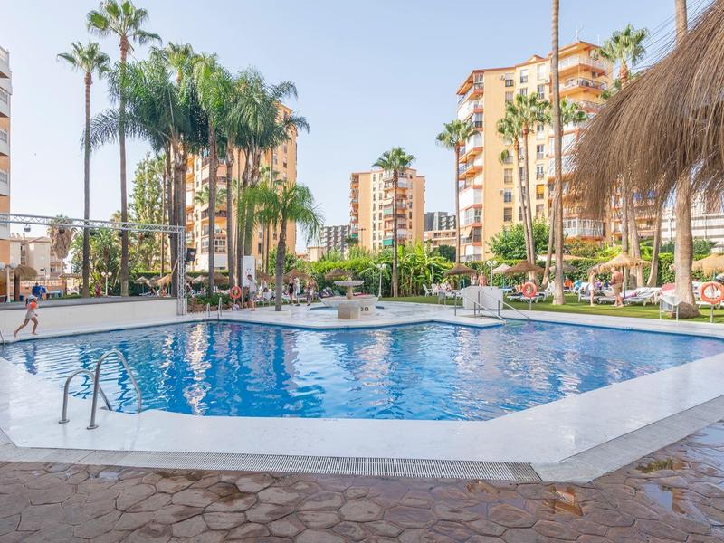 Large hotel pool with sun umbrellas, palm trees, and surrounding multi-story buildings under clear sky.