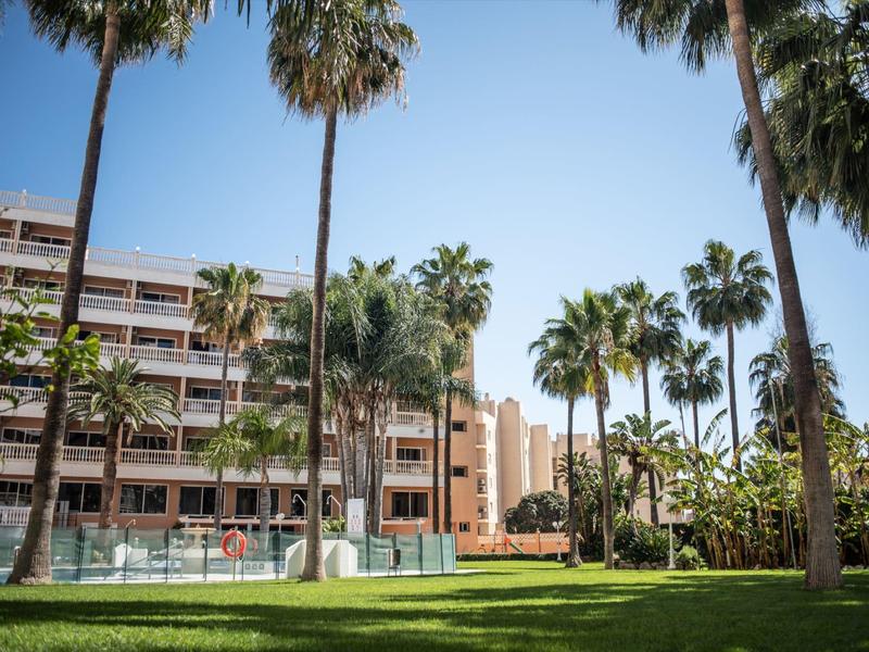 Modern hotel complex with palm trees and large pool under clear blue sky.