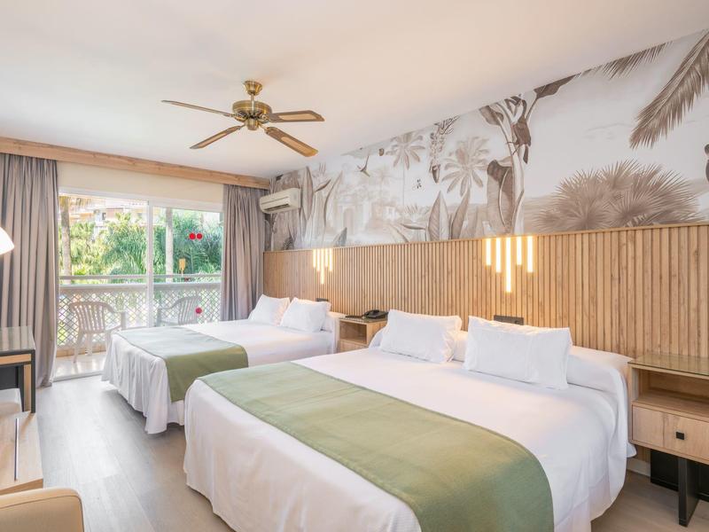 Bright hotel room with two beds, wooden wall, ceiling fan, and balcony overlooking greenery.