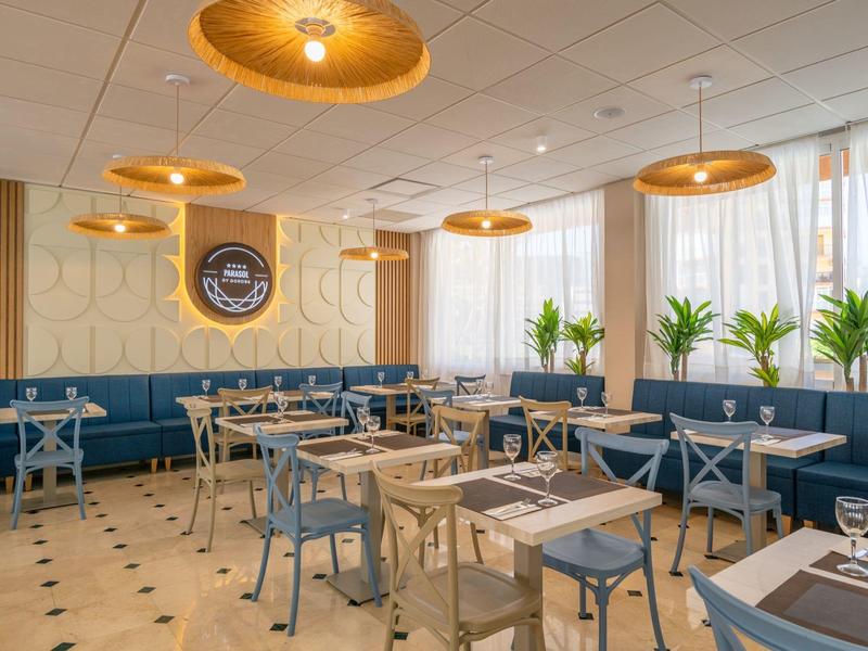 Modern restaurant with blue chairs, wooden tables, and large windows with plants.