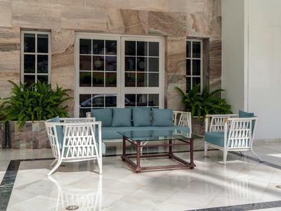 Area salotto esterna moderna con sedie bianche, divano blu, tavolo in vetro e legno, piante e grandi finestre.