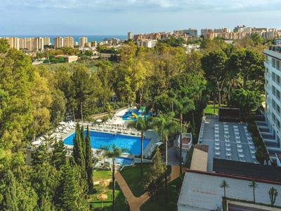 Blick auf ein Hotel mit Pool, umgeben von Bäumen und einer Stadt im Hintergrund unter blauem Himmel.