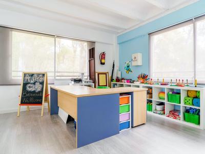 Stanza gioco luminosa con grandi finestre, contenitori colorati e tavolo attività in legno.