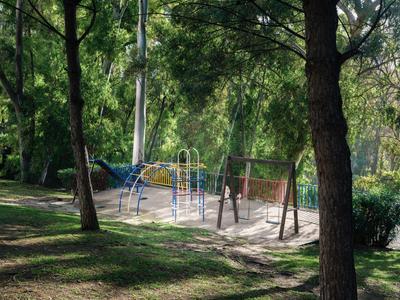 Kleiner Spielplatz mit Schaukeln und Klettergerüst, umgeben von grünen Bäumen in einem Park.