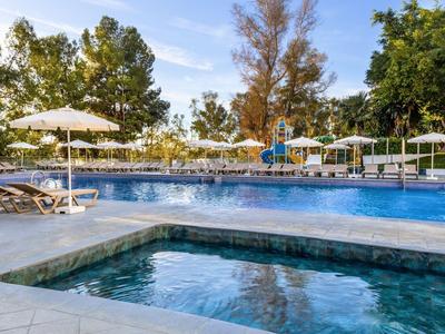 Poolbereich mit Liegestühlen und Sonnenschirmen in einer von Bäumen umgebenen Hotelanlage.
