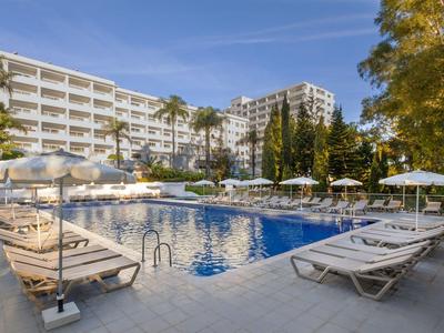 Moderner Hotelpool mit weißen Liegestühlen und Sonnenschirmen, umgeben von Gebäuden und Bäumen.