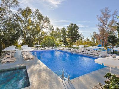 Ein großer rechteckiger Pool mit weißen Sonnenschirmen und Liegestühlen in einer von Bäumen umgebenen Hotelanlage.