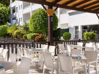 Freiluftrestaurant mit weißen Stühlen und Tischen vor einem Hotelgebäude und gepflegtem Garten.