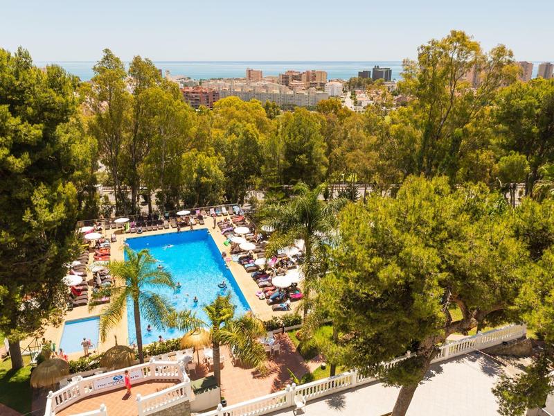 Piscina del resort circondata da alberi e lettini con città e mare sullo sfondo.