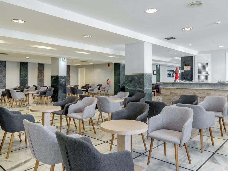 Lounge moderno in hotel con poltrone grigie e tavoli in legno su pavimento decorato.