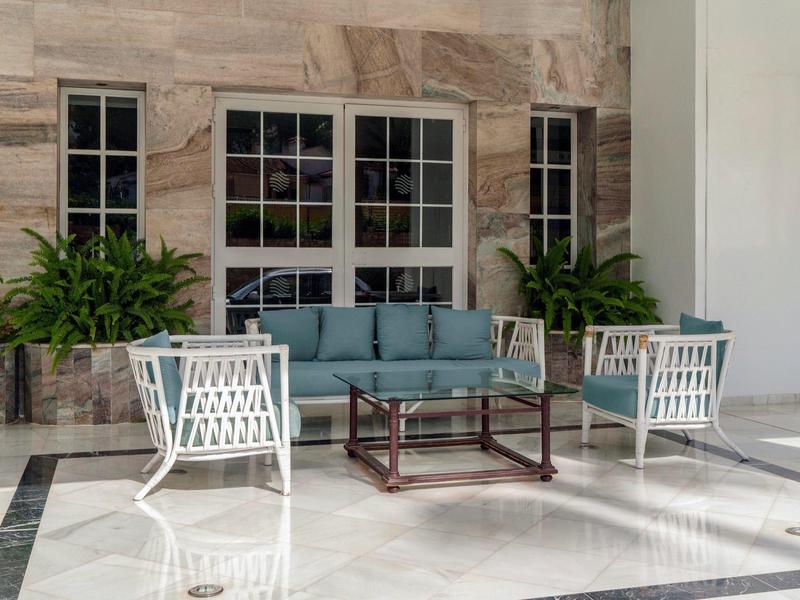 Area salotto esterna moderna con sedie bianche, divano blu, tavolo in vetro e legno, piante e grandi finestre.