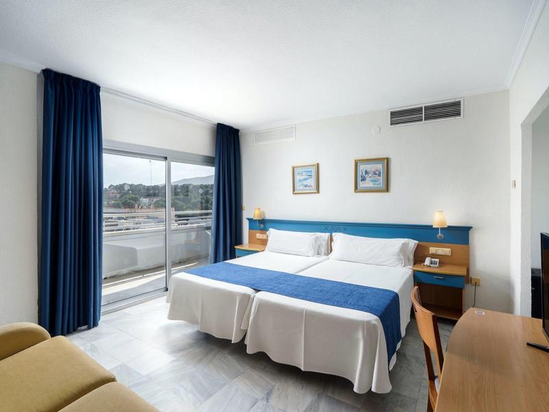 Camera d'hotel luminosa con due letti singoli, tende blu e balcone con vista sulla città.