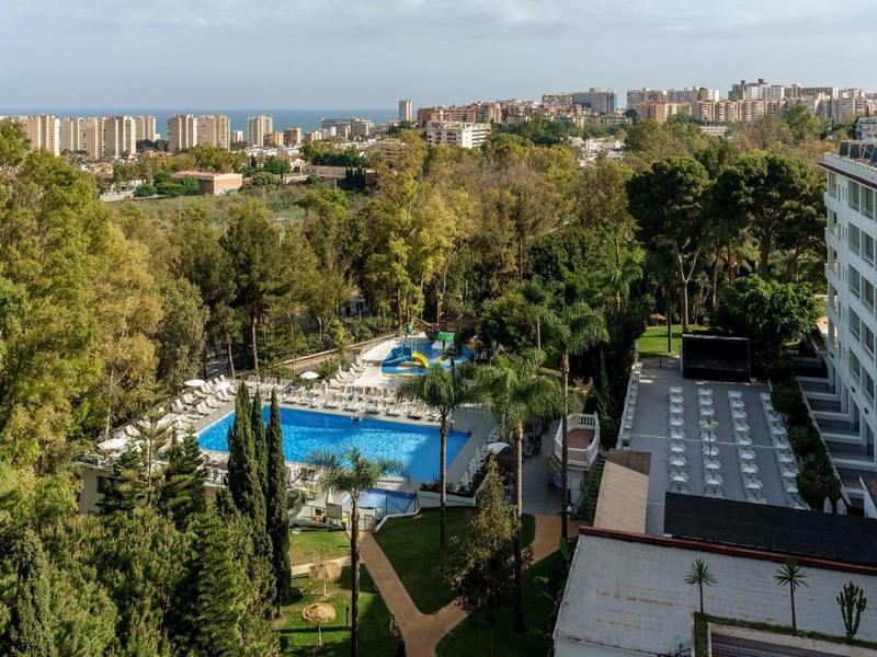 Vista di un hotel con piscina e giardino circondato da alberi e panorama urbano in lontananza.