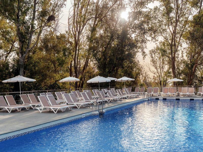 Piscina all'aperto con sdraio e ombrelloni circondata da alberi al sole.