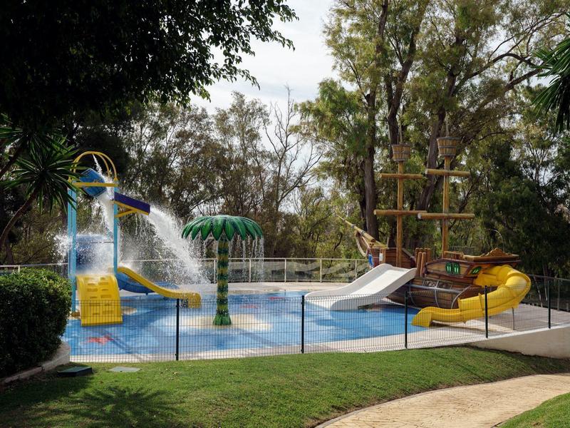 Area giochi acquatica esterna con scivoli e giochi d'acqua circondata da alberi e recinzione.