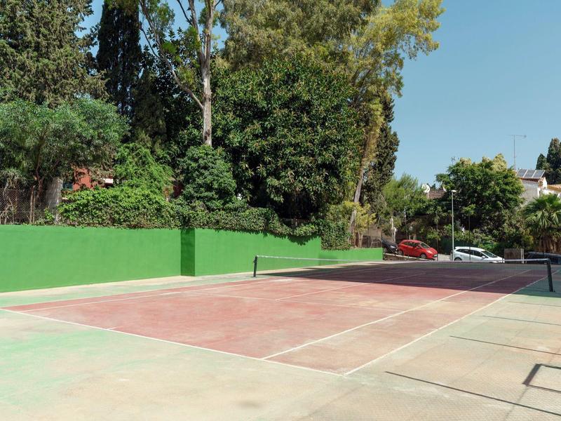 Campo da tennis all'aperto vuoto con recinzione verde e alberi sotto il cielo sereno.