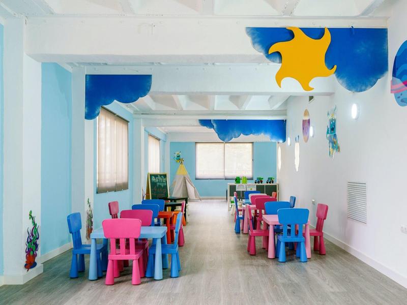 Sala giochi luminosa per bambini con tavoli e sedie colorati sotto decorazioni divertenti al soffitto.