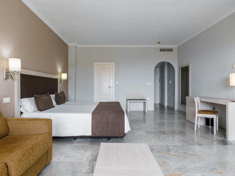 Ampia camera d'albergo con letto grande, divano, scrivania e decorazioni minimaliste in tonalità neutre.