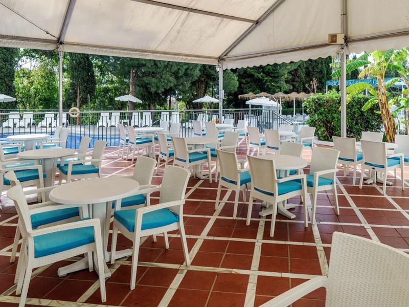 Zona pranzo all'aperto dell'hotel con tavoli bianchi e sedie blu sotto una tettoia su pavimento piastrellato.