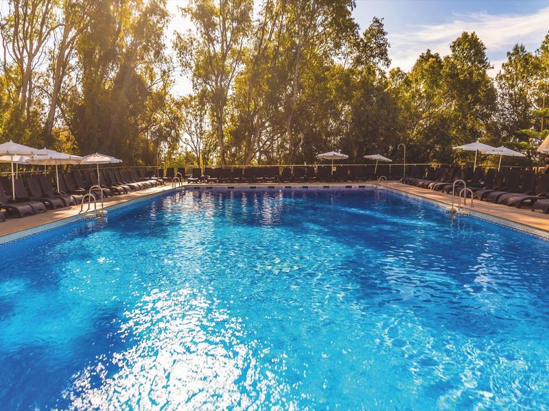 Piscina esterna circondata da lettini e alberi al sole.