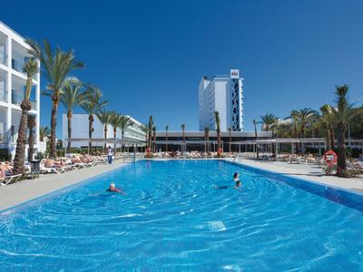 Großer Hotelpool mit Sonnenliegen, Palmen und blauem Himmel an einem sonnigen Tag.