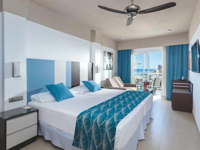 Camera d'hotel moderna con letto matrimoniale, ventilatore a soffitto e balcone con vista mare.