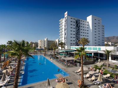 Moderner Hotelkomplex mit großem blauen Pool, Palmen und Liegestühlen bei sonnigem Wetter.