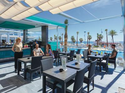 Moderner Außenbereich eines Hotels mit Sitzplätzen, Pool und Palmen unter klarem Himmel.