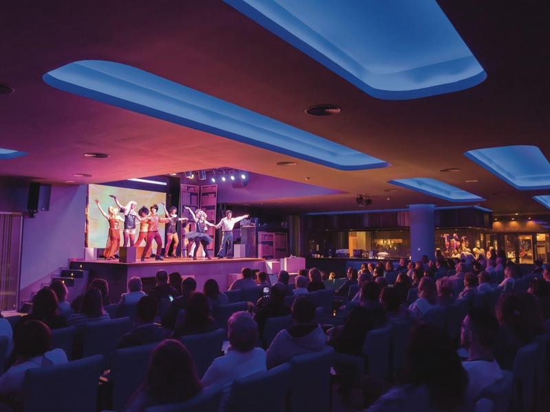 Spettacolo su palco con pubblico seduto in teatro con luci blu e viola.