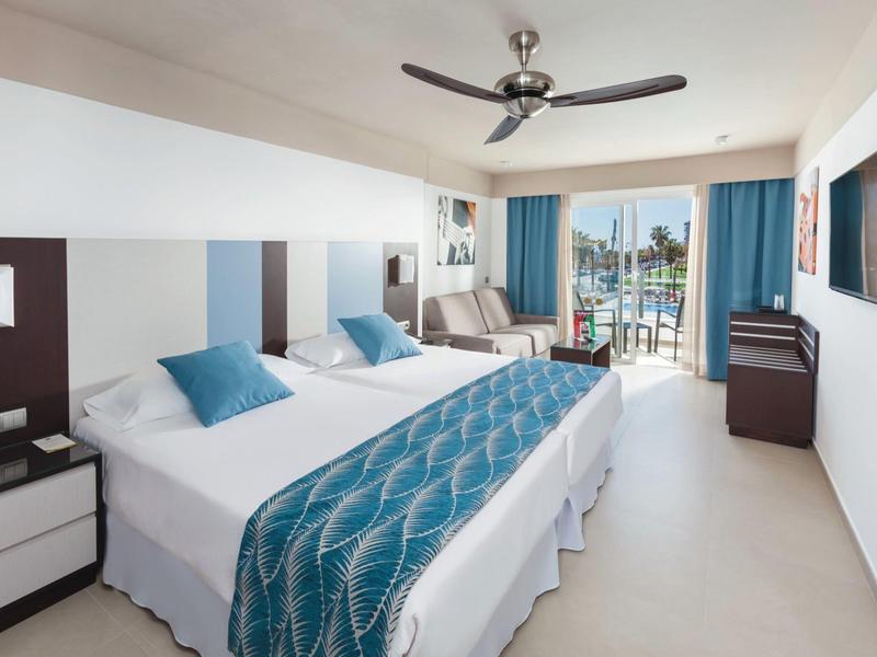 Camera d'hotel moderna con due letti, decorazioni blu e balcone con vista sul mare.