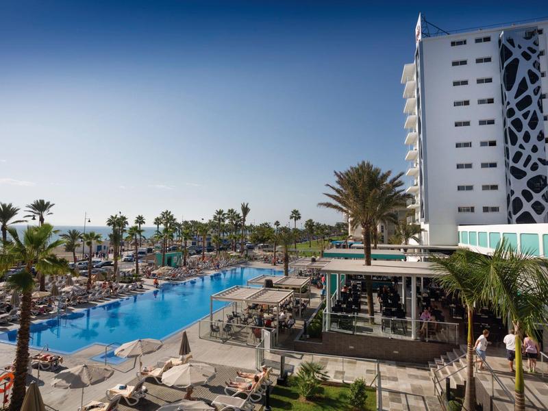 Piscina all'aperto con palme e lettini accanto a un moderno hotel bianco sotto un cielo azzurro.