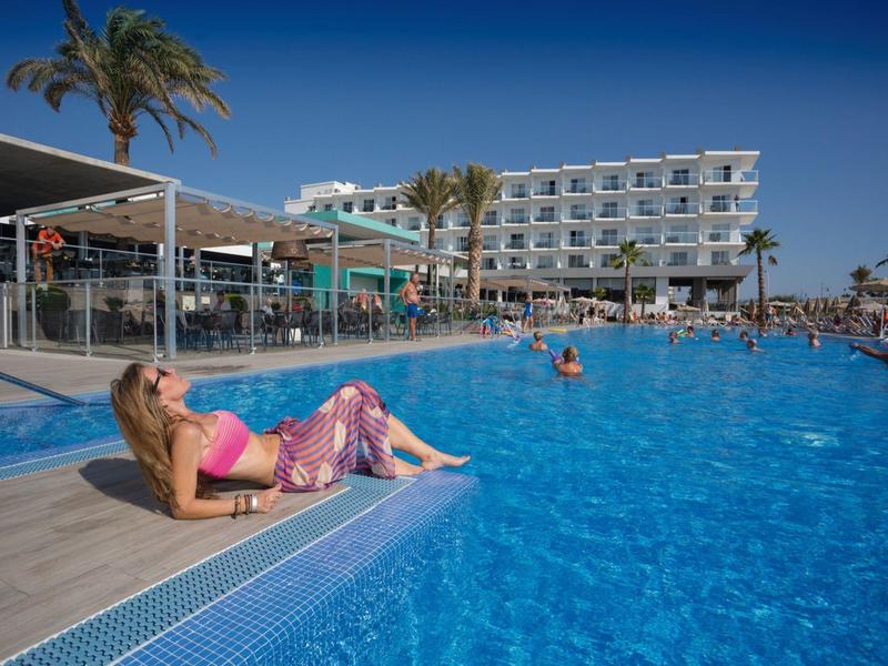 Donna rilassata a bordo piscina di un hotel con cielo limpido e palme.