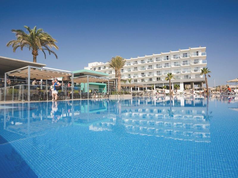 Grande piscina limpida davanti a un hotel moderno sotto un cielo blu e una palma.