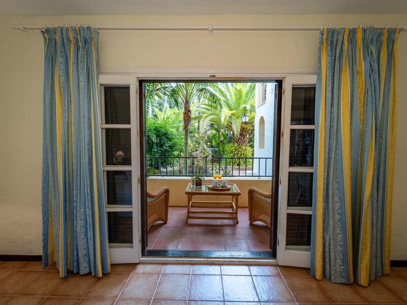 Vue à travers des portes vitrées ouvertes sur une terrasse avec une table en bois et des plantes tropicales.