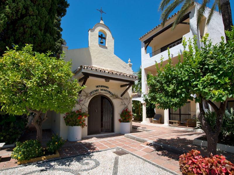 Petite chapelle à côté du bâtiment de l'hôtel avec des arbres et un ciel ensoleillé.