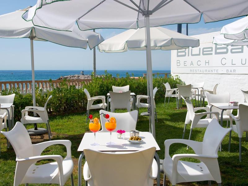 Terrasse d'un club de plage avec des chaises blanches, des tables, des parasols et vue sur la mer.
